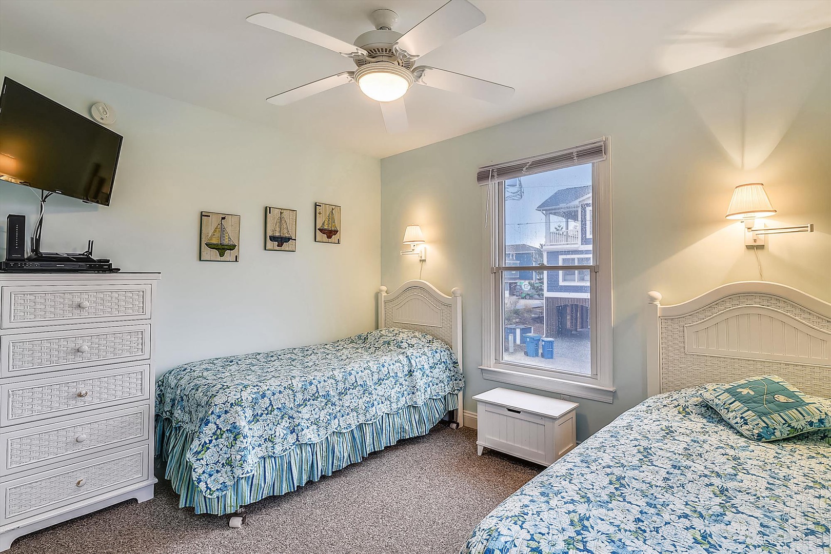 Your bright bedroom features two comfortable beds with coastal-style linens, ceiling fan for cool breezes, flat-screen TV, dresser storage, and bedside lighting for reading.