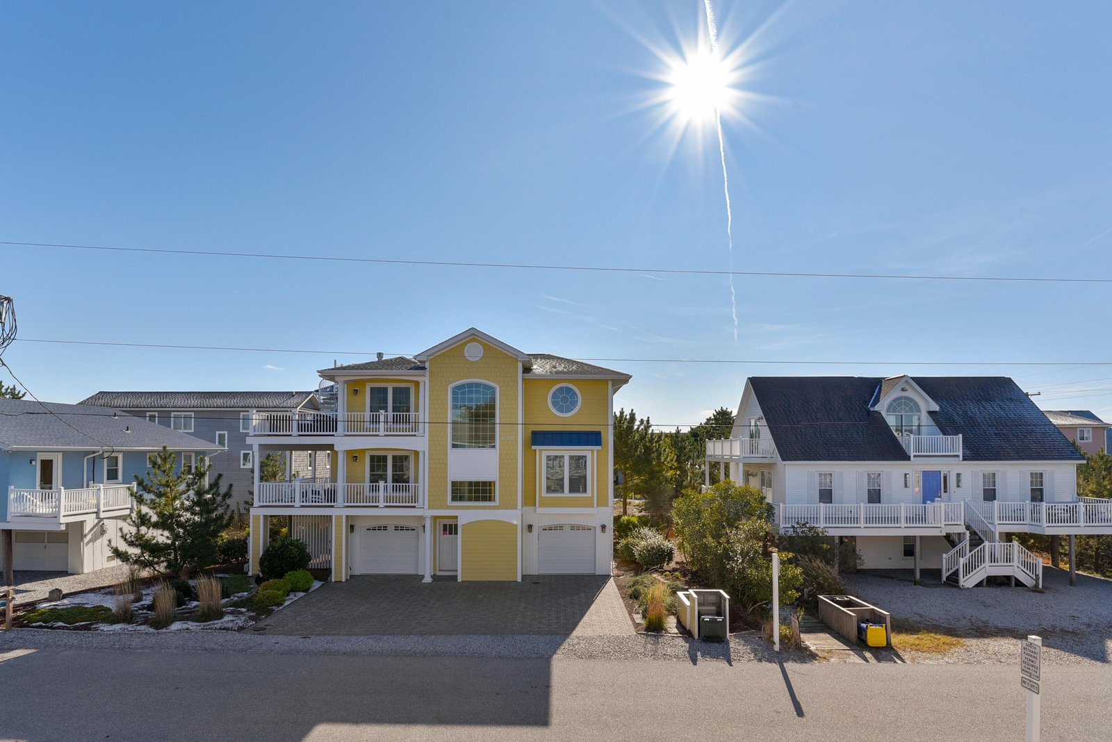 A charming yellow coastal home with white trim and distinctive round windows, surrounded by neighboring beach houses in a quiet residential neighborhood.