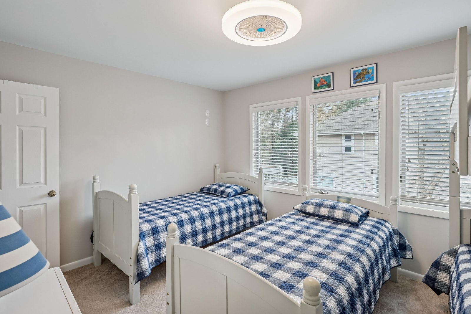 Rest easy in this bright, airy bedroom featuring twin beds with cozy blue-checkered bedding. Natural light pours in through large windows, and a ceiling fan keeps you comfortable.