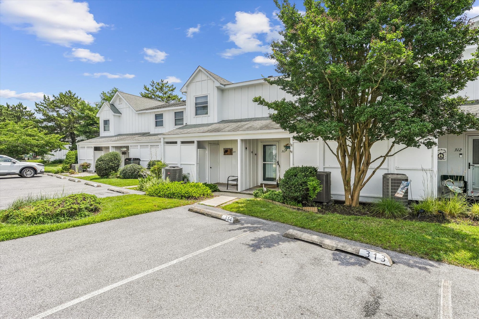 Modern white-siding townhomes with private garages, set in a peaceful suburban neighborhood with mature trees and landscaped green spaces framing the entrance.