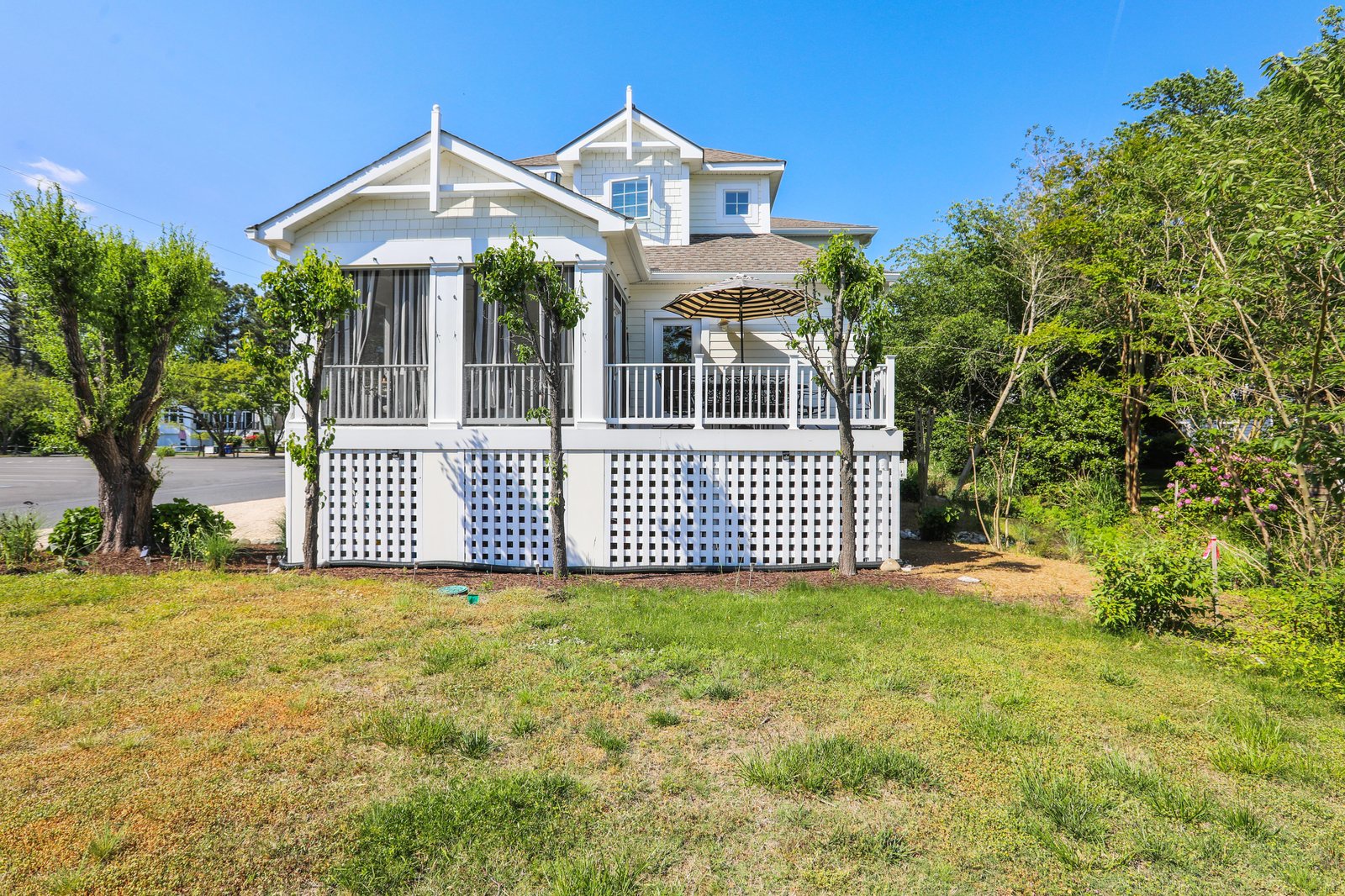 This charming two-story coastal cottage features an elevated wrap-around porch, lattice skirting, and a shaded outdoor seating area, surrounded by mature trees and natural greenery.