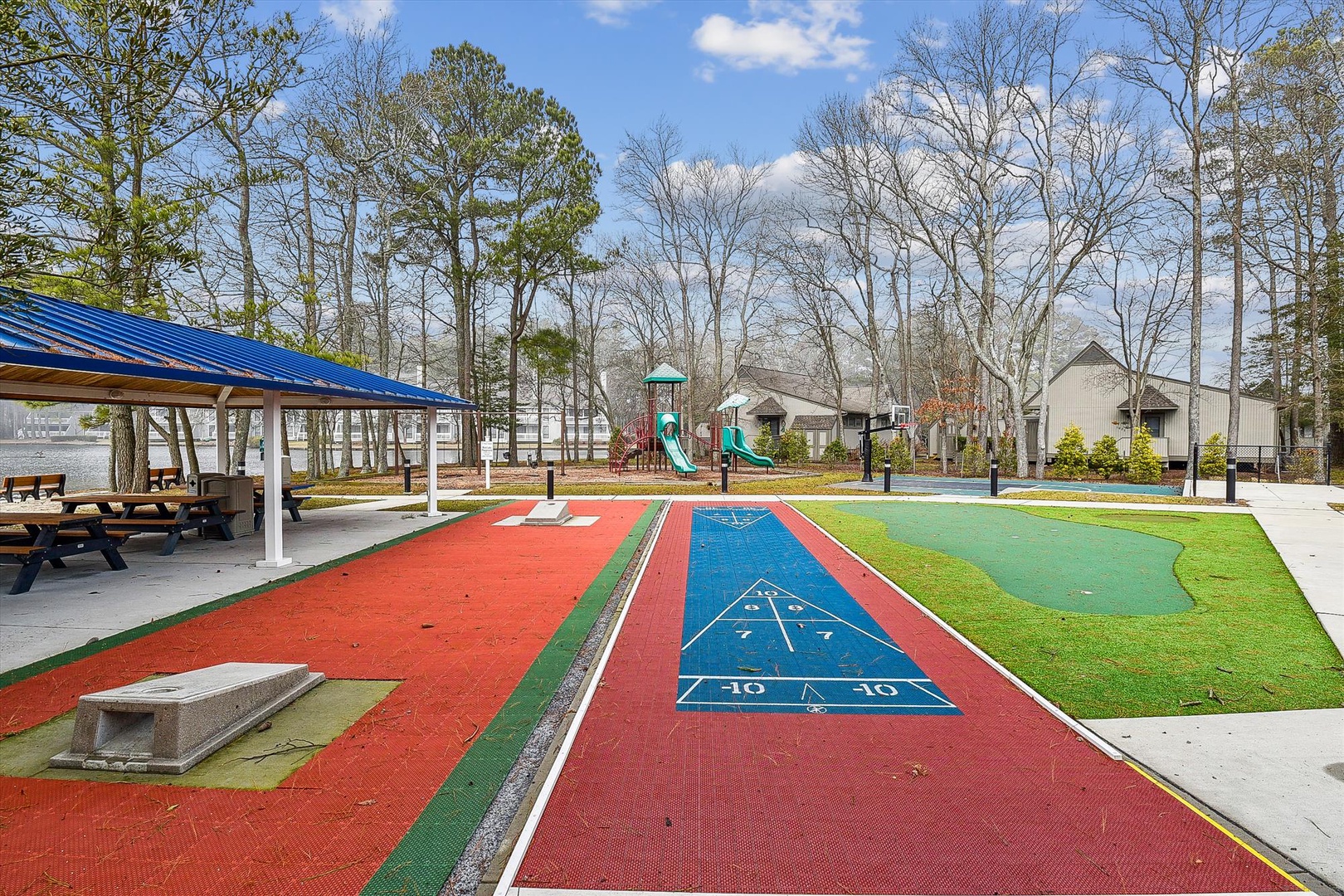 A vibrant outdoor recreation area featuring shuffleboard courts, picnic pavilion, and playground facilities surrounded by towering trees and residential buildings.
