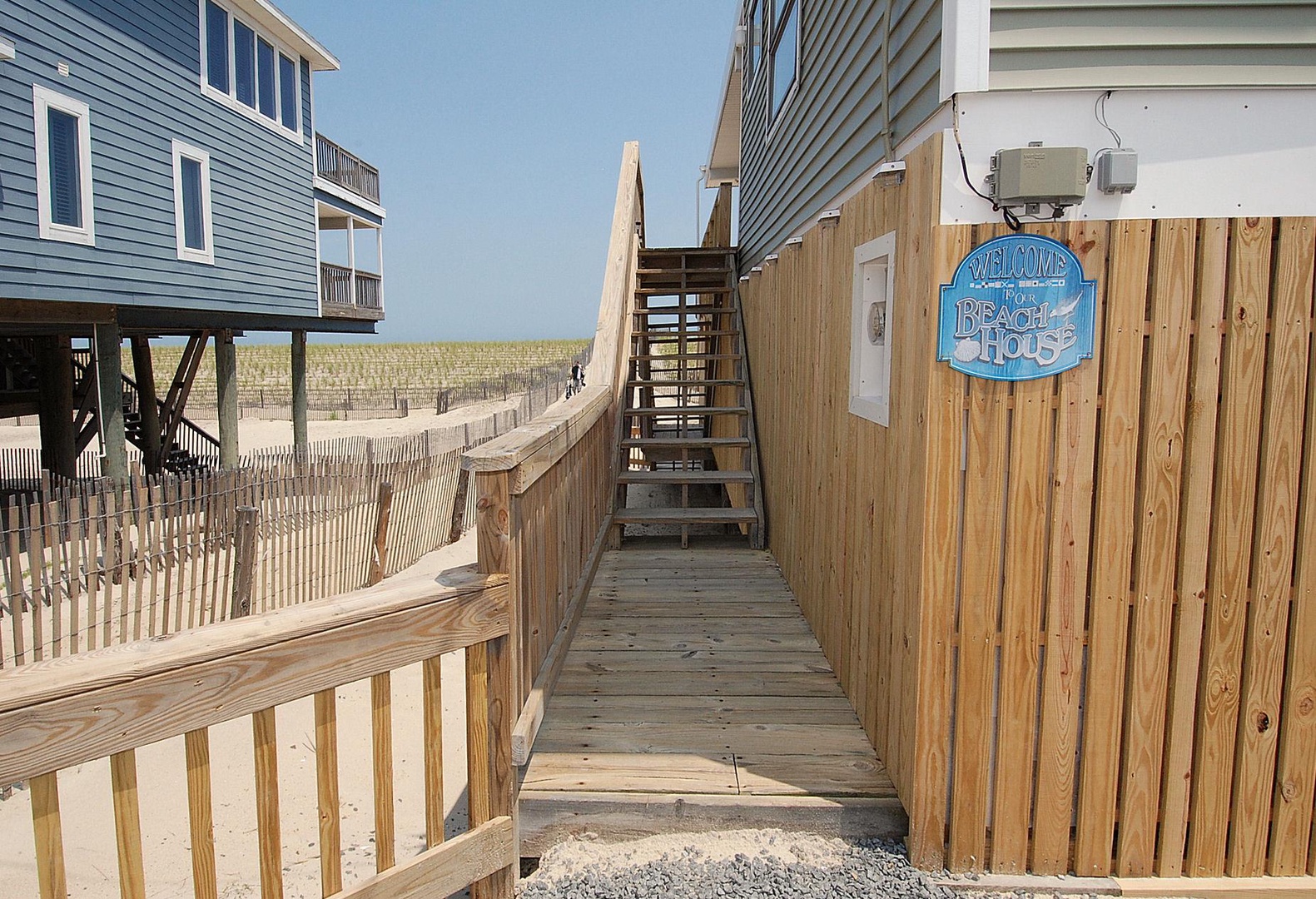 A welcoming boardwalk path leads through coastal homes to the beach, marked by a cheerful 'Beach House' sign at the entrance. Sand dunes and fencing frame the journey to the shore.