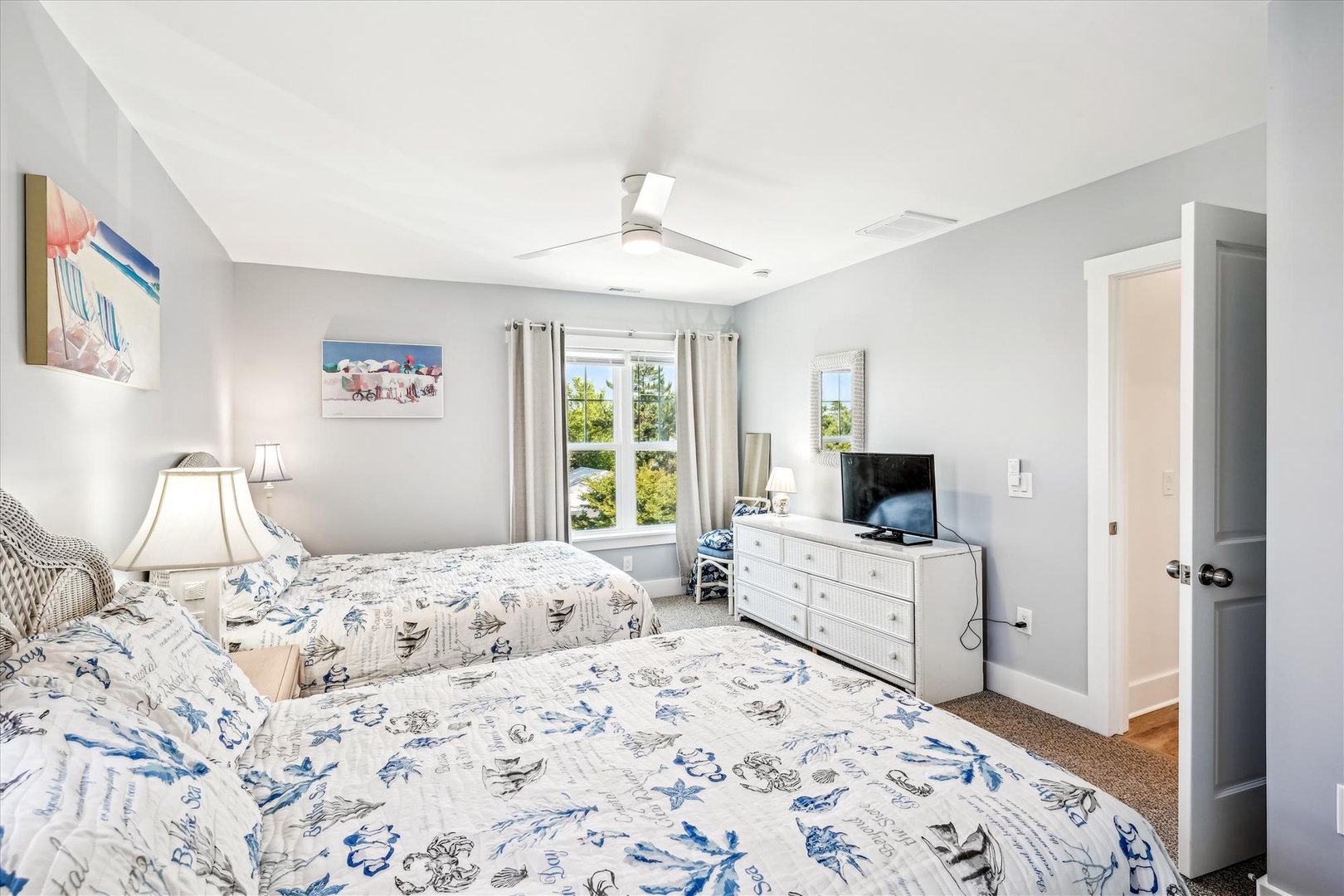 Wake up in your bright, coastal-themed bedroom with two comfortable beds, natural light streaming through windows, and beach artwork setting the perfect seaside mood.