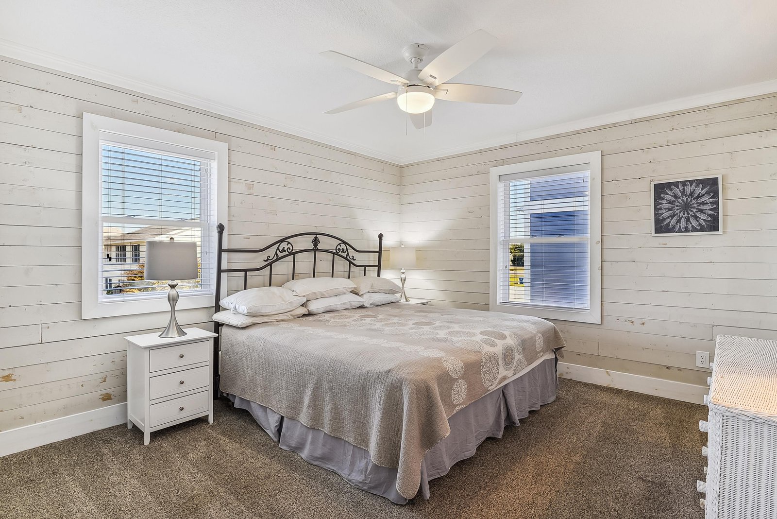 Drift off in this serene bedroom with whitewashed shiplap walls and a cozy coastal feel, where gentle breezes from the ceiling fan and soft natural light through the windows create your perfect retreat.