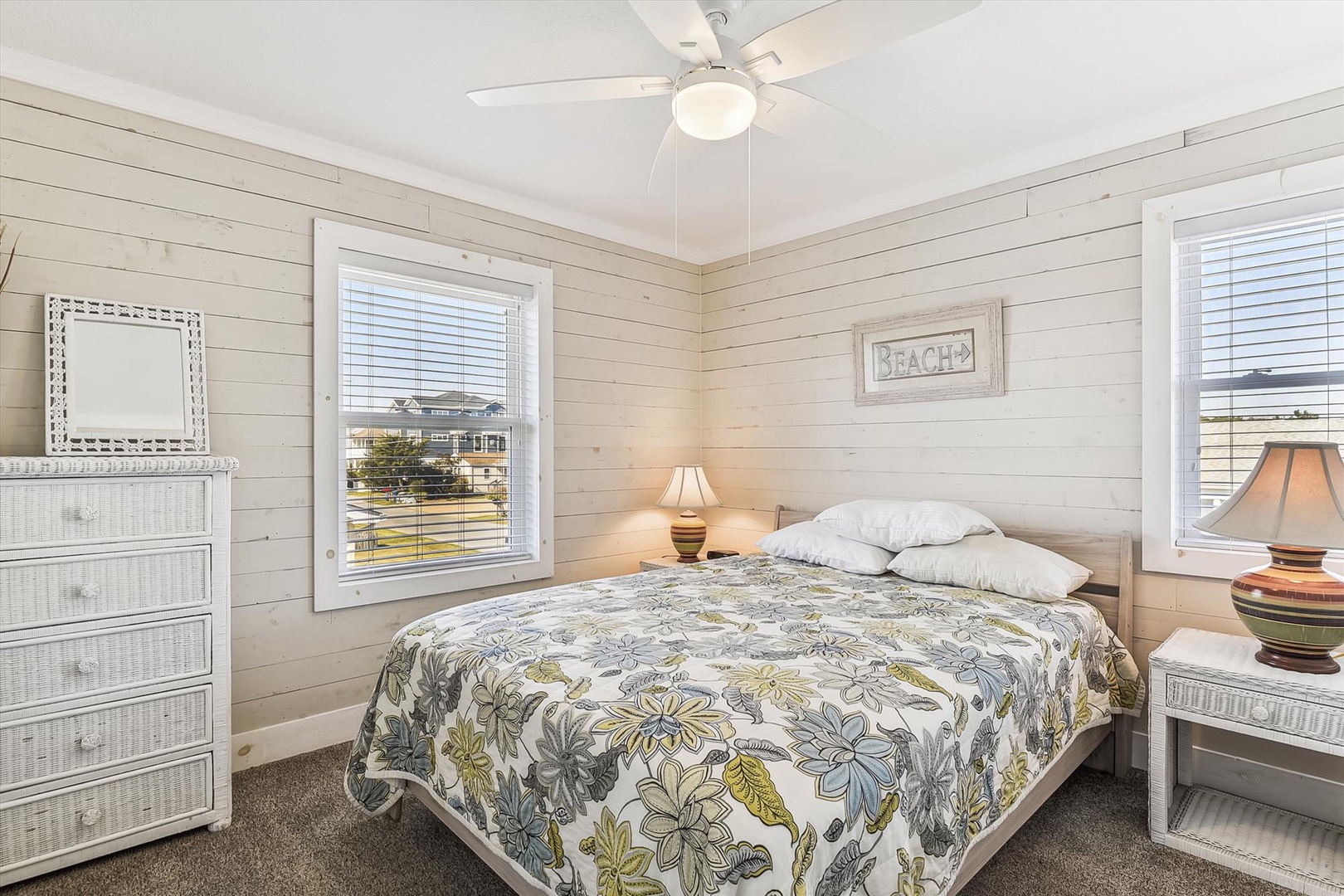 Unwind in your cozy coastal bedroom featuring shiplap walls, soft bedding, and two windows that frame peaceful neighborhood views. A ceiling fan and beach-inspired décor complete your restful retreat.
