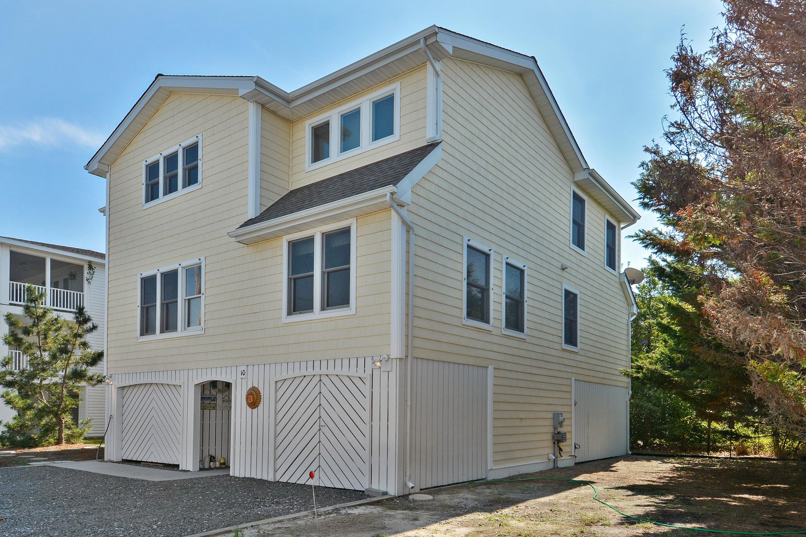 This charming three-story beach house features classic coastal architecture with light siding and ample windows, nestled in a serene neighborhood surrounded by mature trees and natural landscaping.