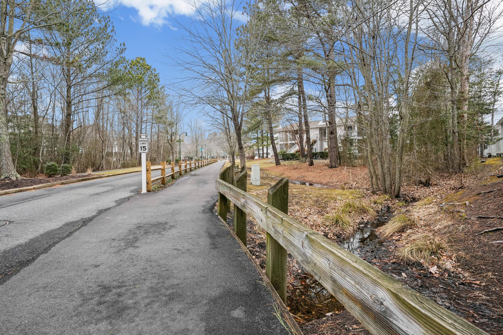 Tree-lined residential street with wooden fencing leads to the property entrance, offering a peaceful neighborhood setting with natural wooded surroundings.
