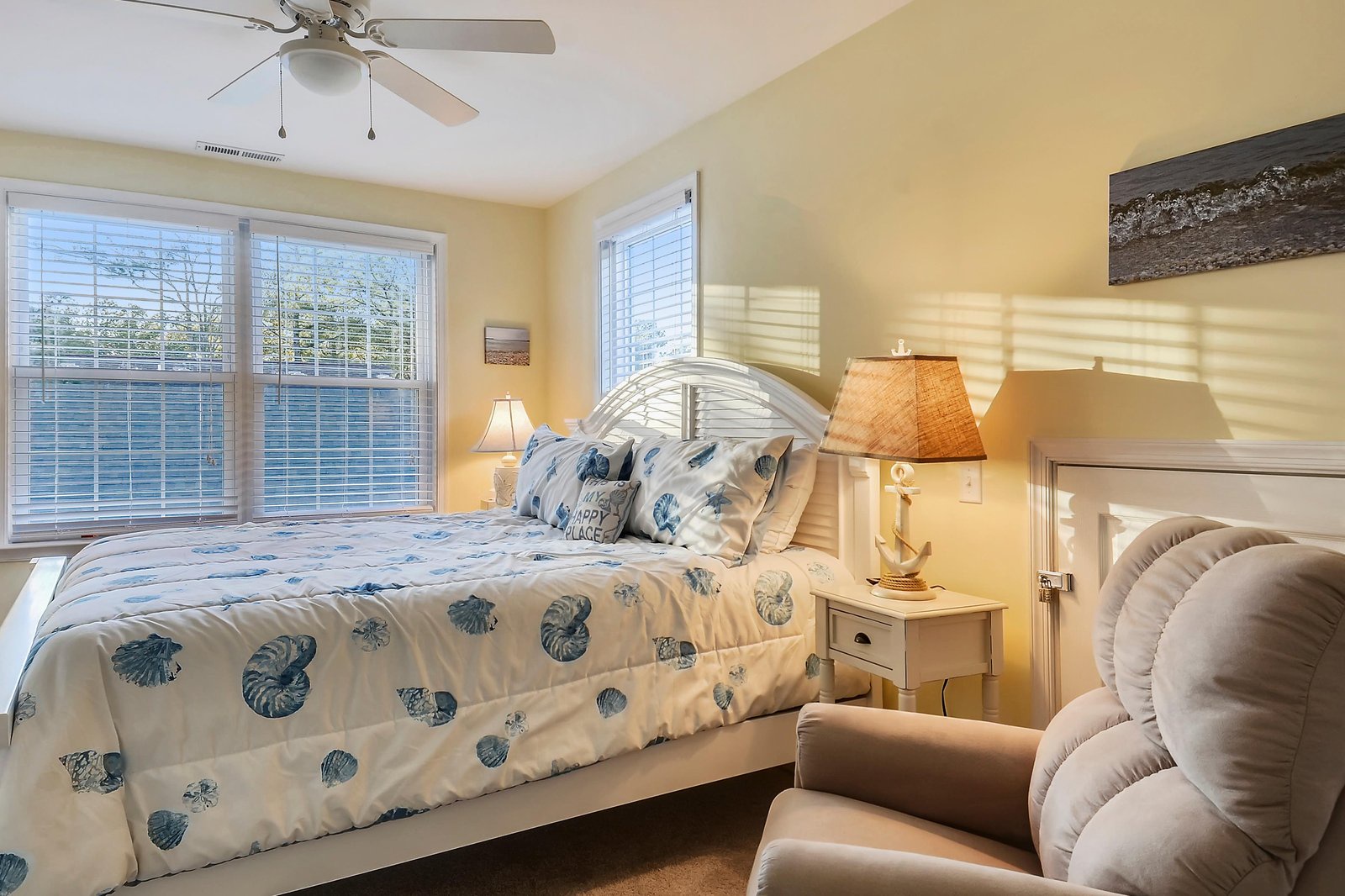 Step into your serene coastal retreat, where soft morning light filters through the windows and a plush bed awaits. Sink into the comfortable chair and feel the gentle breeze from the ceiling fan—this is your peaceful haven by the shore.