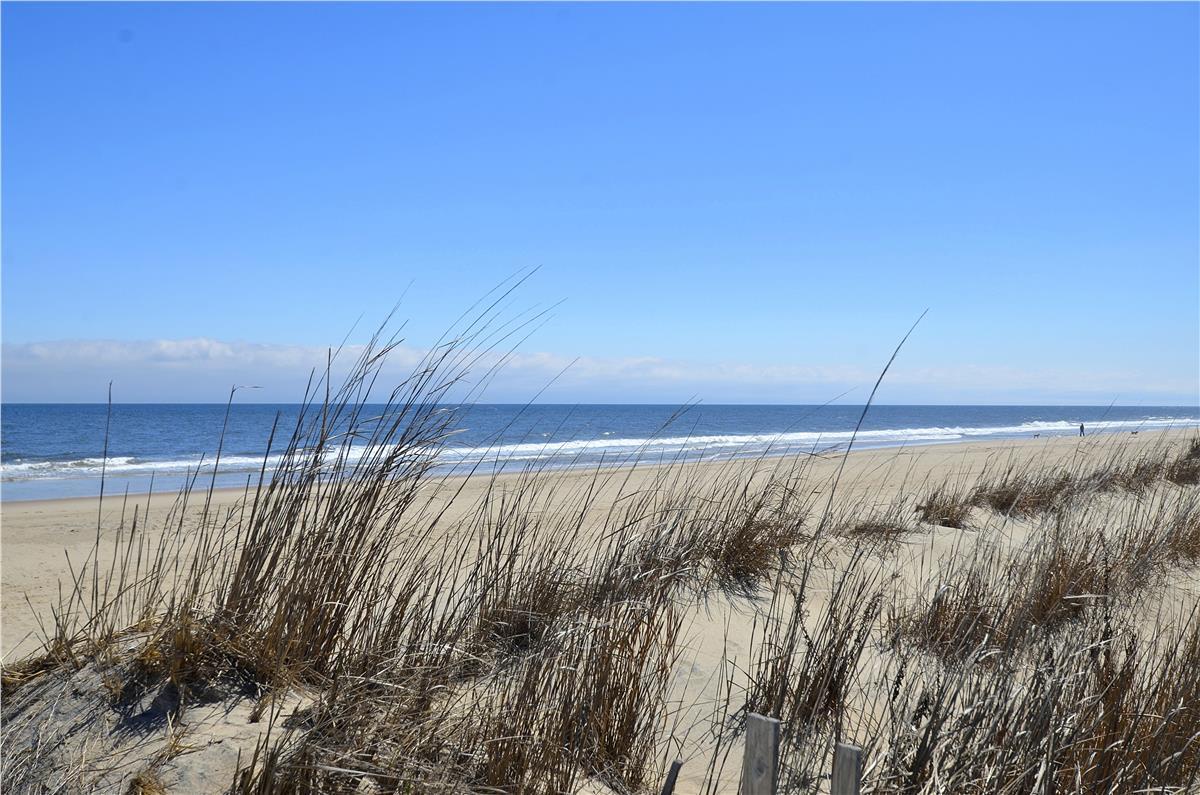 A pristine beach stretches along the coast, framed by dunes and sea grass, with gentle waves rolling onto soft sand under clear skies.