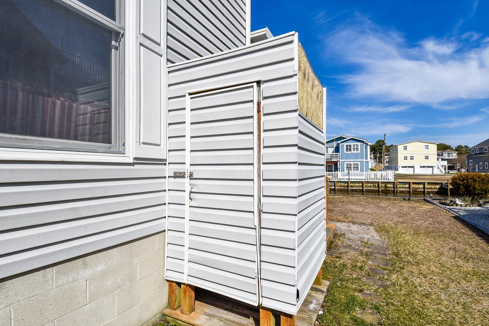 The property features a private outdoor space surrounded by neighboring coastal homes, with clear blue skies overhead and a quiet residential setting.