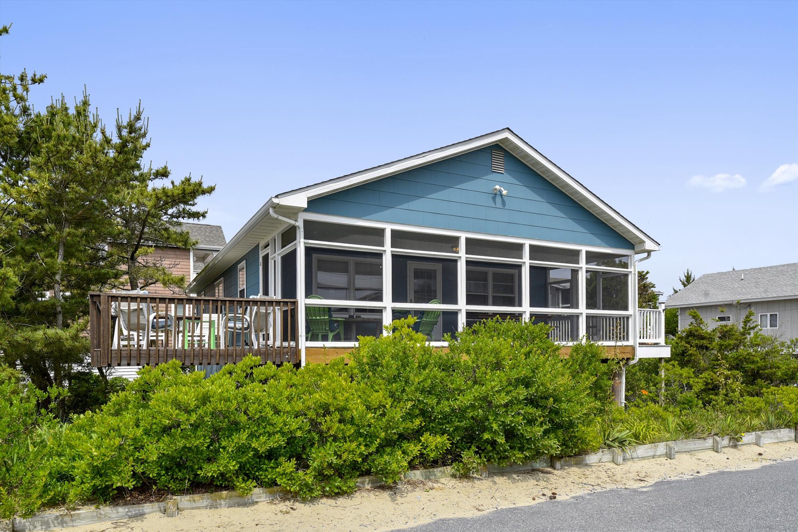 Charming blue beach house with screened porch and wooden deck, nestled among coastal greenery with nearby homes visible in the area.