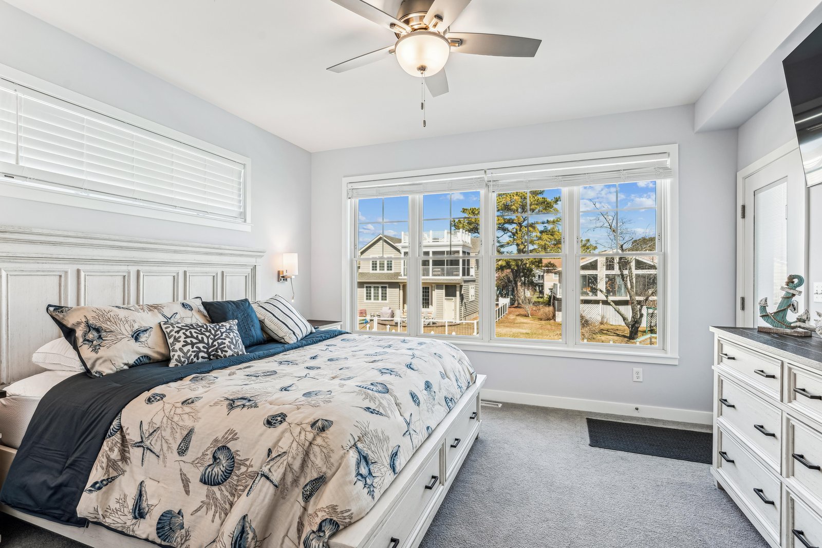 Relax in this inviting coastal bedroom with soft linens and a ceiling fan. Natural light pours through large windows, offering peaceful neighborhood views.