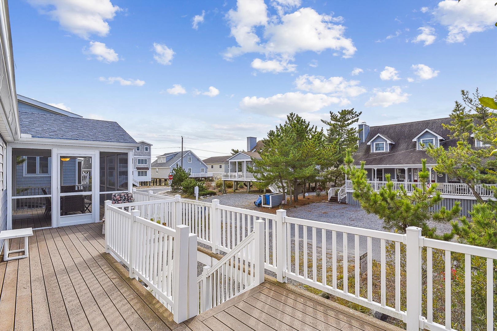 A coastal neighborhood setting with beach homes visible beyond a spacious wooden deck and white railing, offering a quiet residential atmosphere near the shore.
