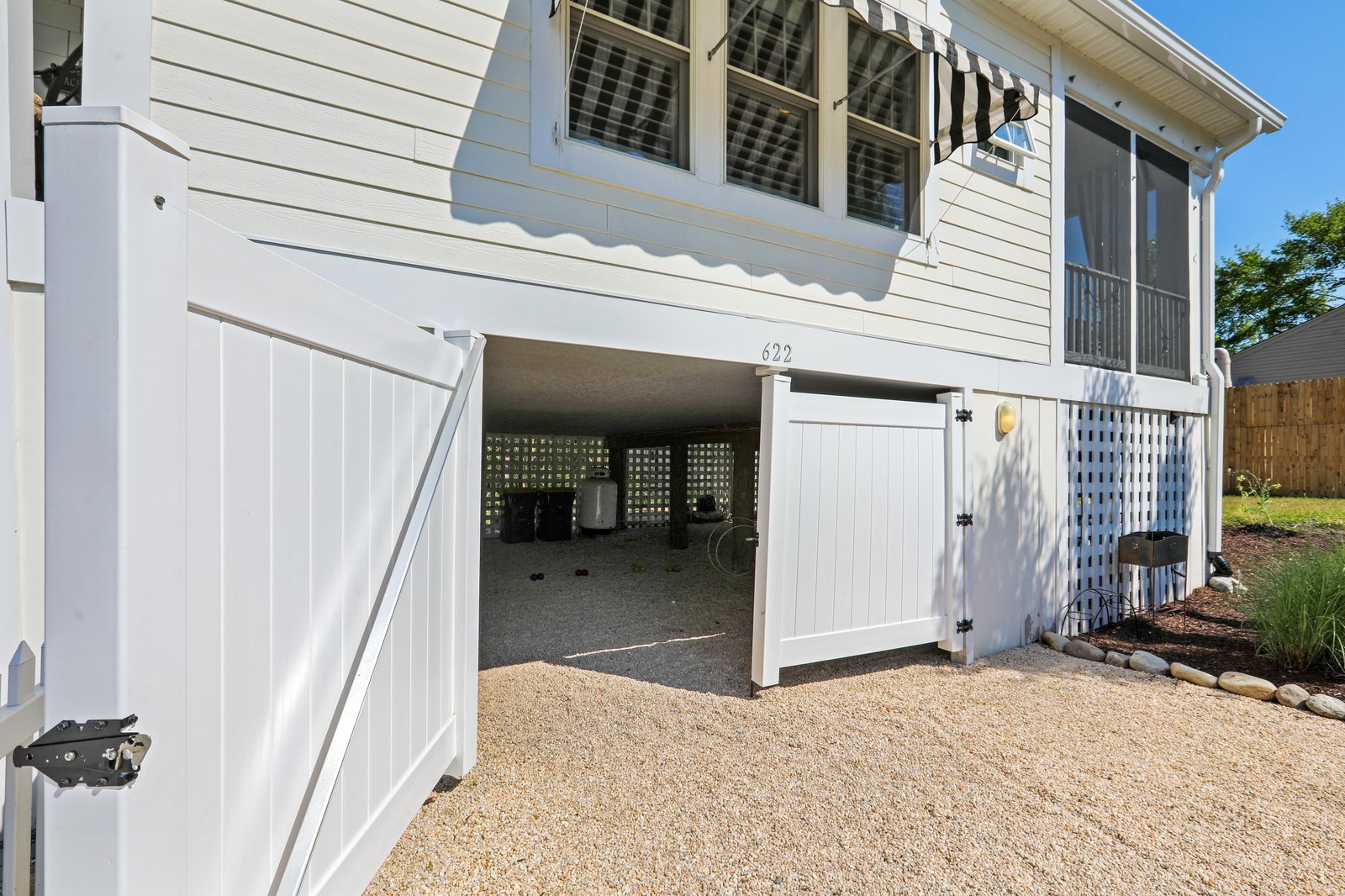 A coastal-style property featuring covered ground-level parking and gated entry, photographed in bright daylight with clear blue skies overhead.