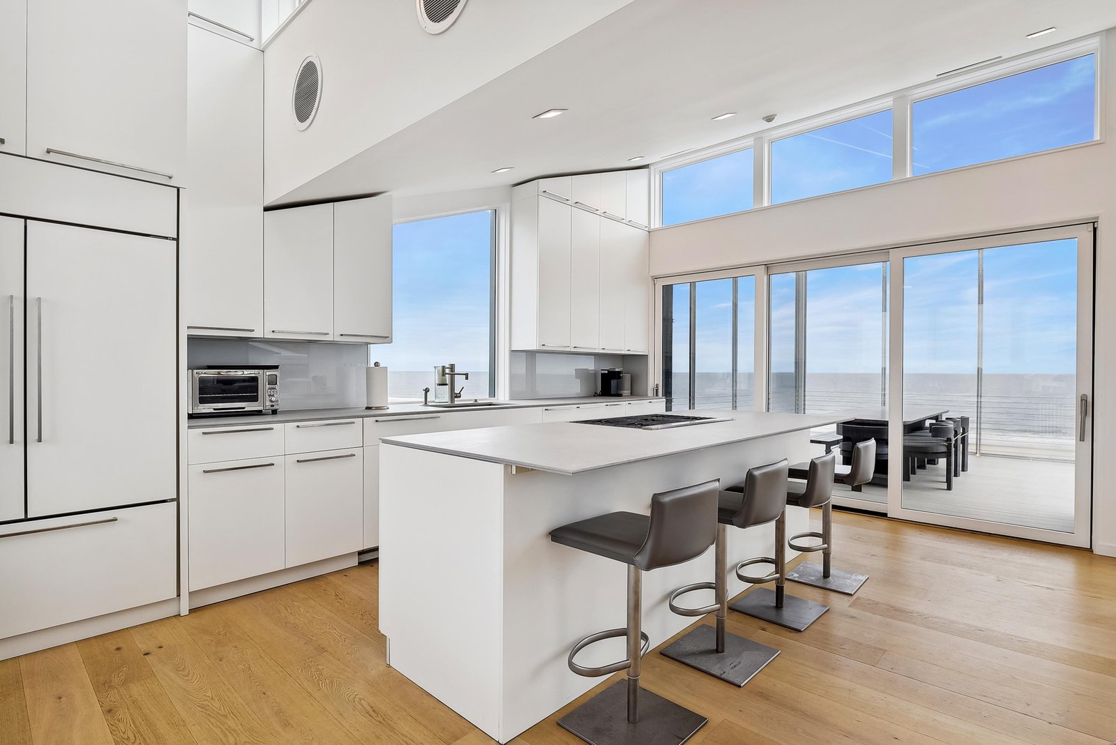 Your stunning modern kitchen welcomes you with sleek white cabinetry, a spacious island with breakfast bar seating, and floor-to-ceiling windows framing peaceful ocean views. Step outside to your private terrace and breathe in the fresh sea air.