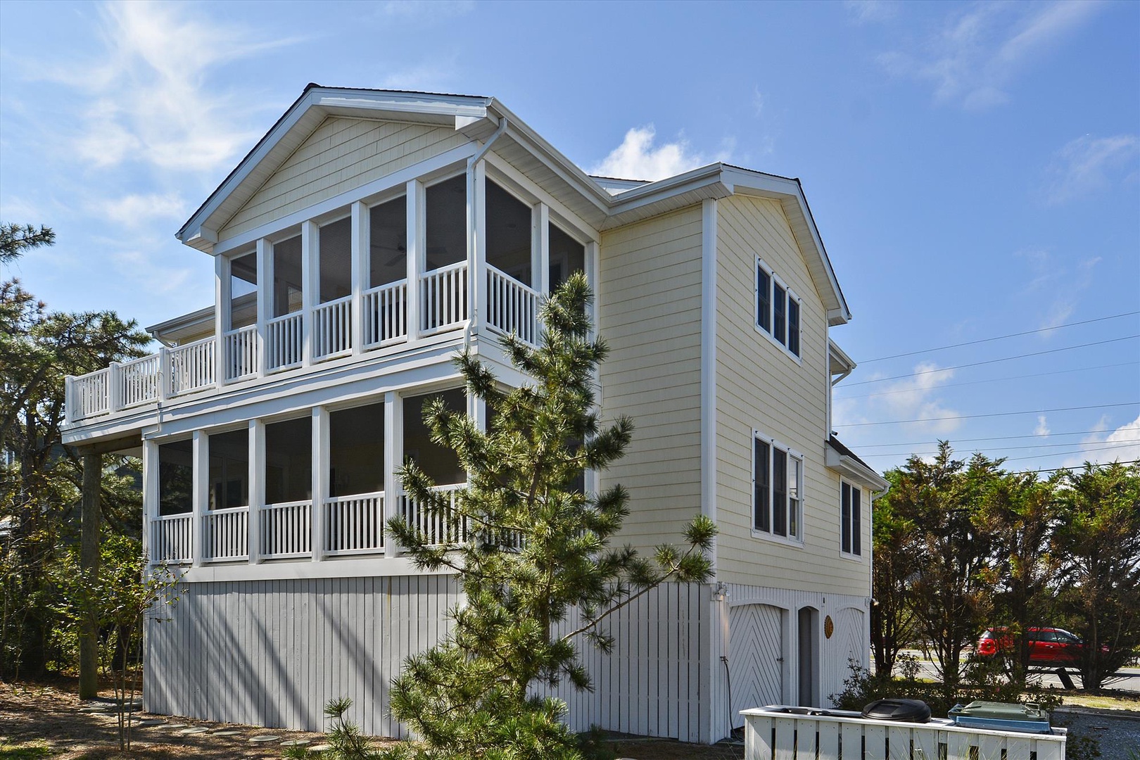 A charming three-story coastal home with wraparound porches and white railings, surrounded by lush greenery under bright blue skies.
