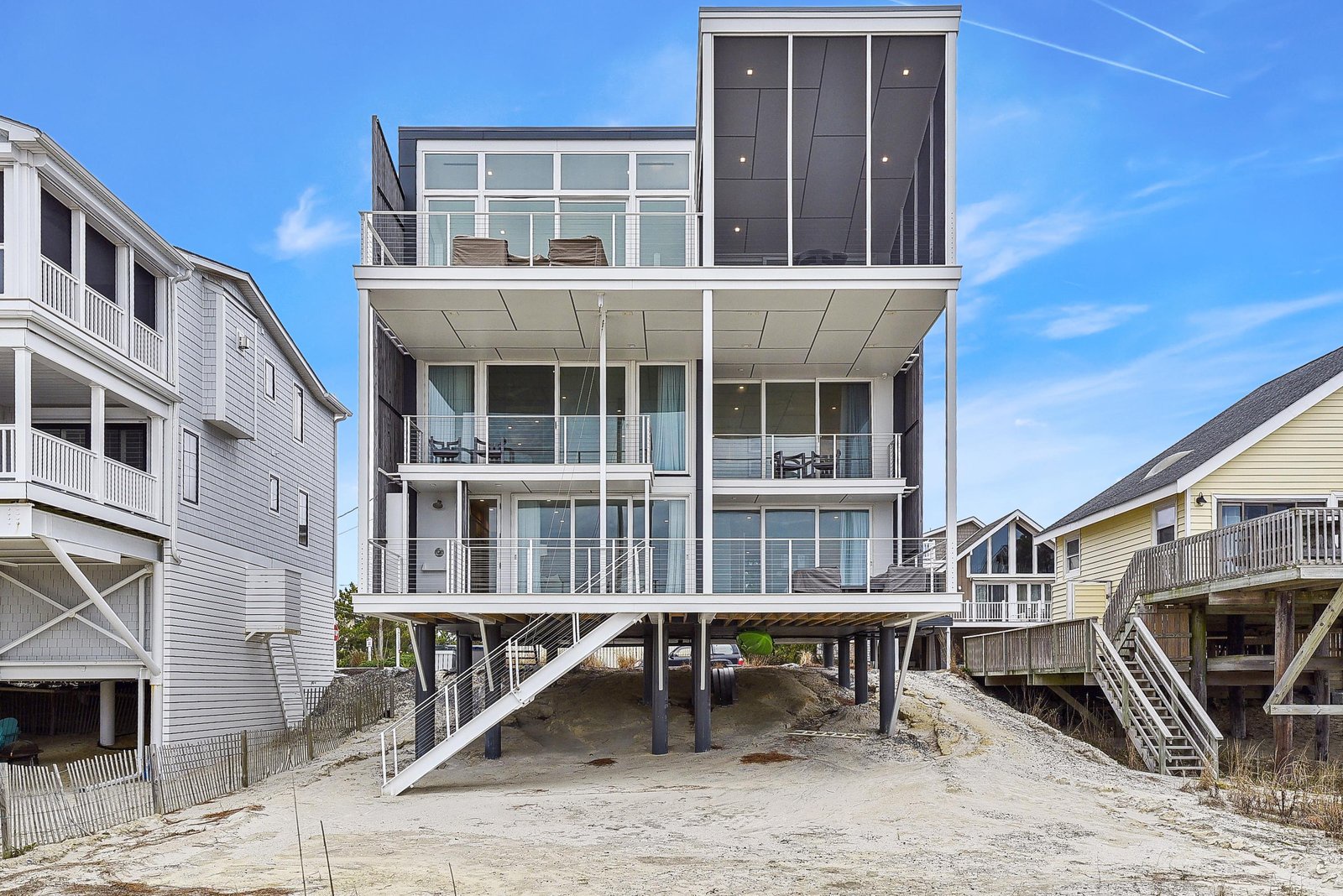 Modern beachfront property with multiple levels of glass-enclosed balconies and direct beach access, nestled in a coastal neighborhood.