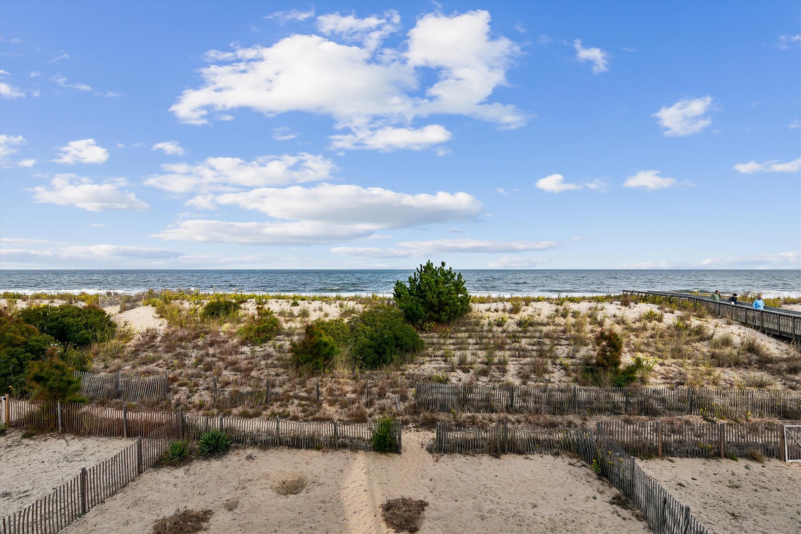 Protected sand dunes bordered by wooden fencing lead to a pristine beach and calm ocean waters under a blue sky with scattered clouds.