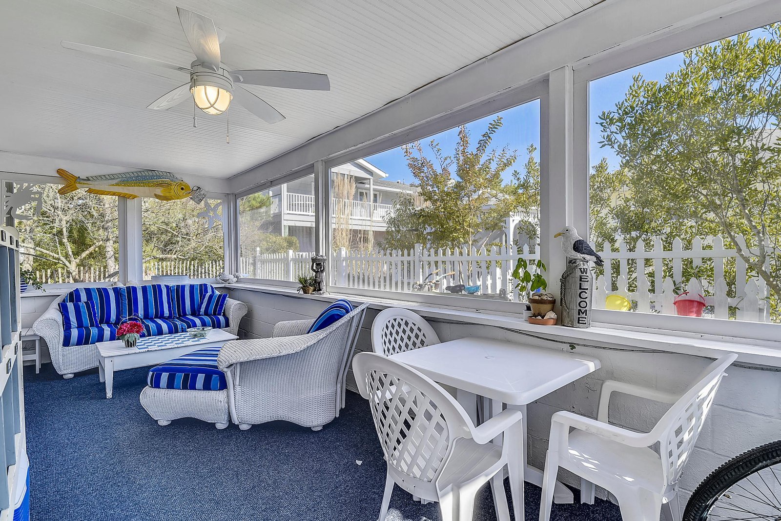 Relax in your screened porch with comfy seating, a bright blue ceiling fan, and a cheerful dining nook. Natural light pours in through wide windows framing greenery and the neighborhood beyond.