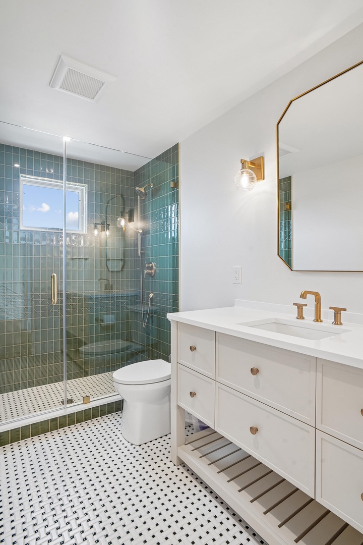 Your private bathroom blends modern style and relaxation, featuring elegant aqua tiles, brass fixtures, and a spacious walk-in shower. Sunlight filters through the window, warming the crisp white vanity and mosaic floor.