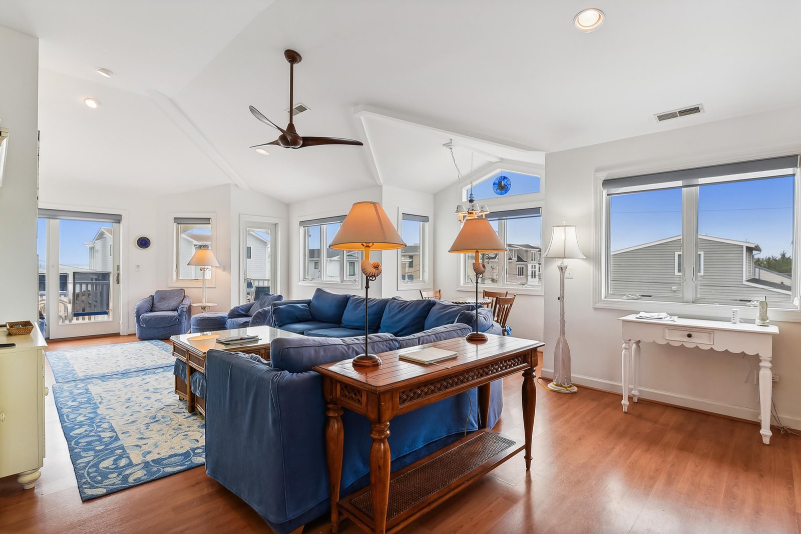 Step into your bright, airy living room where coastal charm meets comfort. Vaulted ceilings and ocean breezes invite you to unwind on plush blue sofas, warmed by natural light pouring through every window.