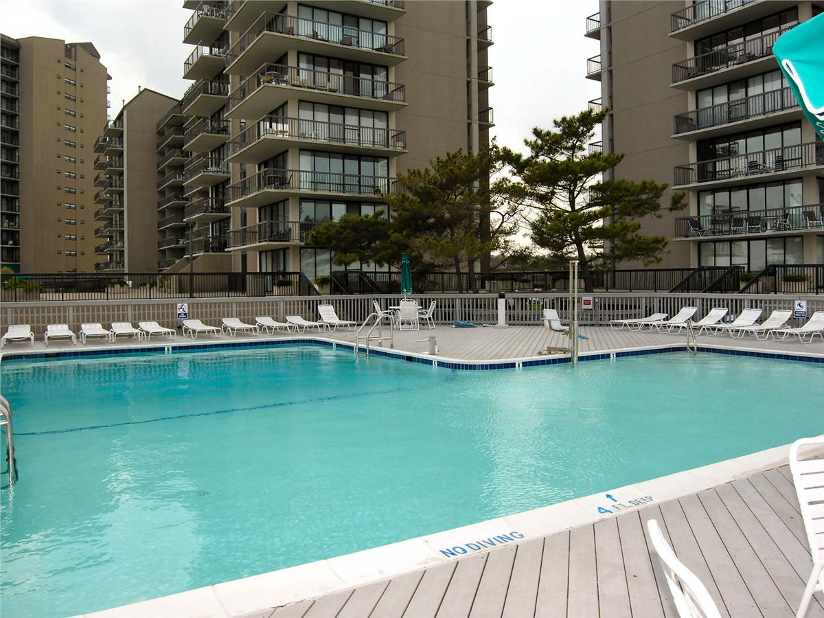 Large outdoor swimming pool surrounded by lounge chairs and pine trees, part of this multi-story beachfront residential complex.