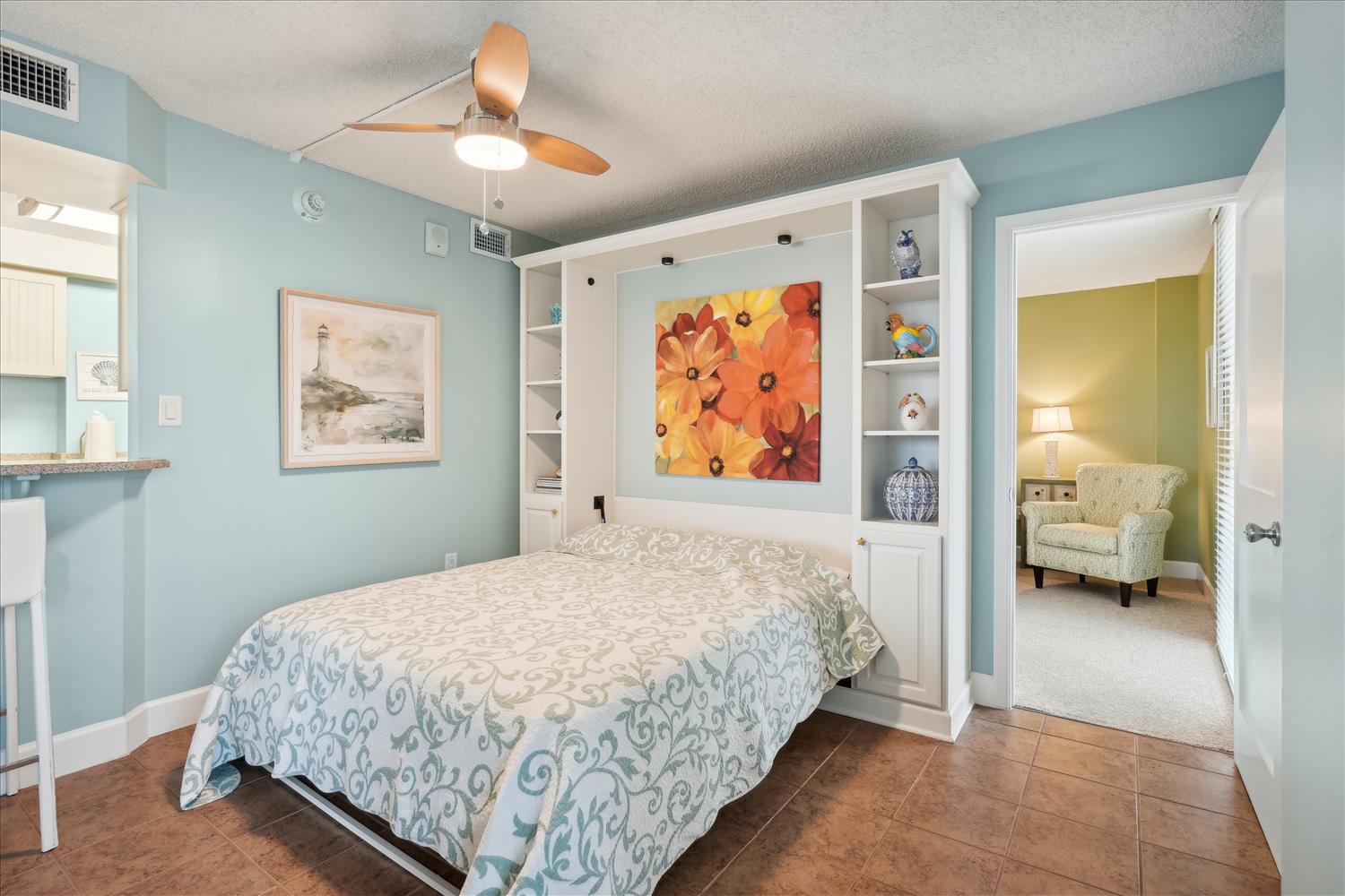 Relax in this serene bedroom with coastal blue walls, a fold-down bed for flexible space, stylish artwork, and a ceiling fan. Your private reading nook awaits just steps away.