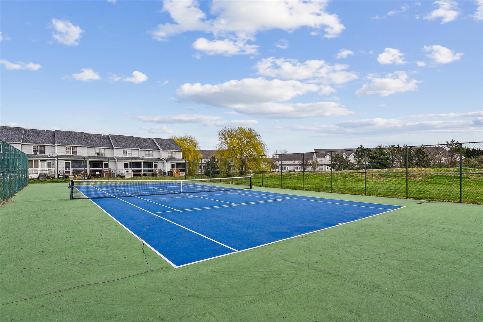 Stay active on the property's private tennis court, featuring a pristine blue hard surface surrounded by open green lawns and peaceful residential views.