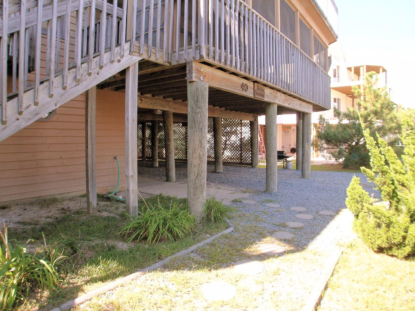 Elevated beachside building featuring covered ground-level parking, outdoor storage, and direct property access via exterior stairway, surrounded by residential neighborhood landscaping.