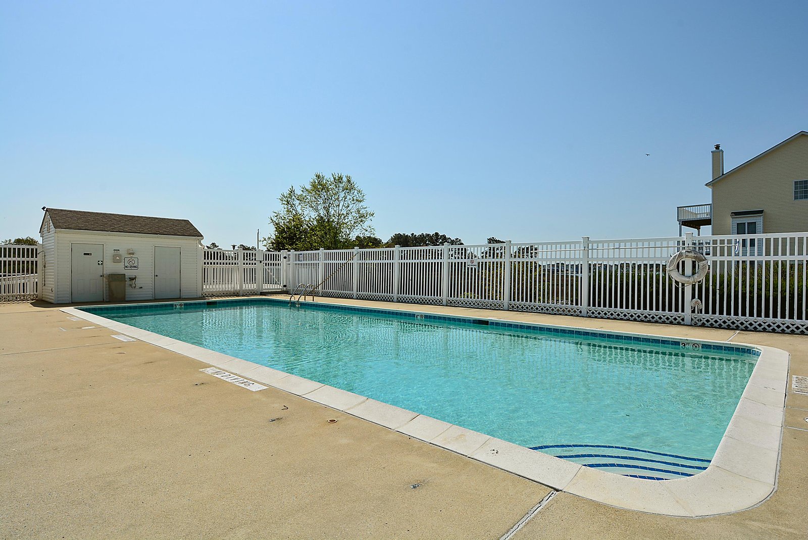 Communal pool area surrounded by white fencing, with ample deck space and clear blue water under sunny skies. Nearby residential buildings offer easy access.