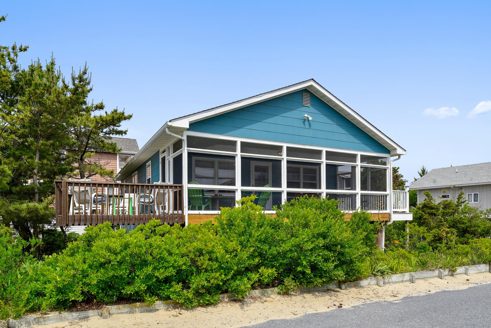 Charming turquoise beach cottage set among dunes and coastal greenery, featuring expansive screened windows and a welcoming deck with seating for relaxation.