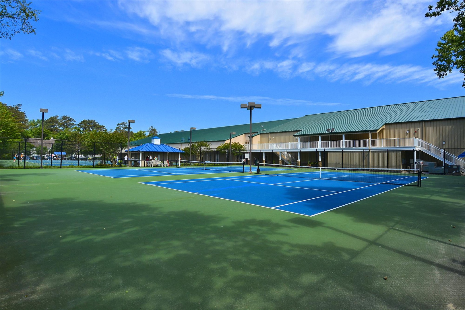 Bright tennis courts with a covered pavilion sit ready for friendly matches, surrounded by mature trees and the property's main building with convenient upper-level access.