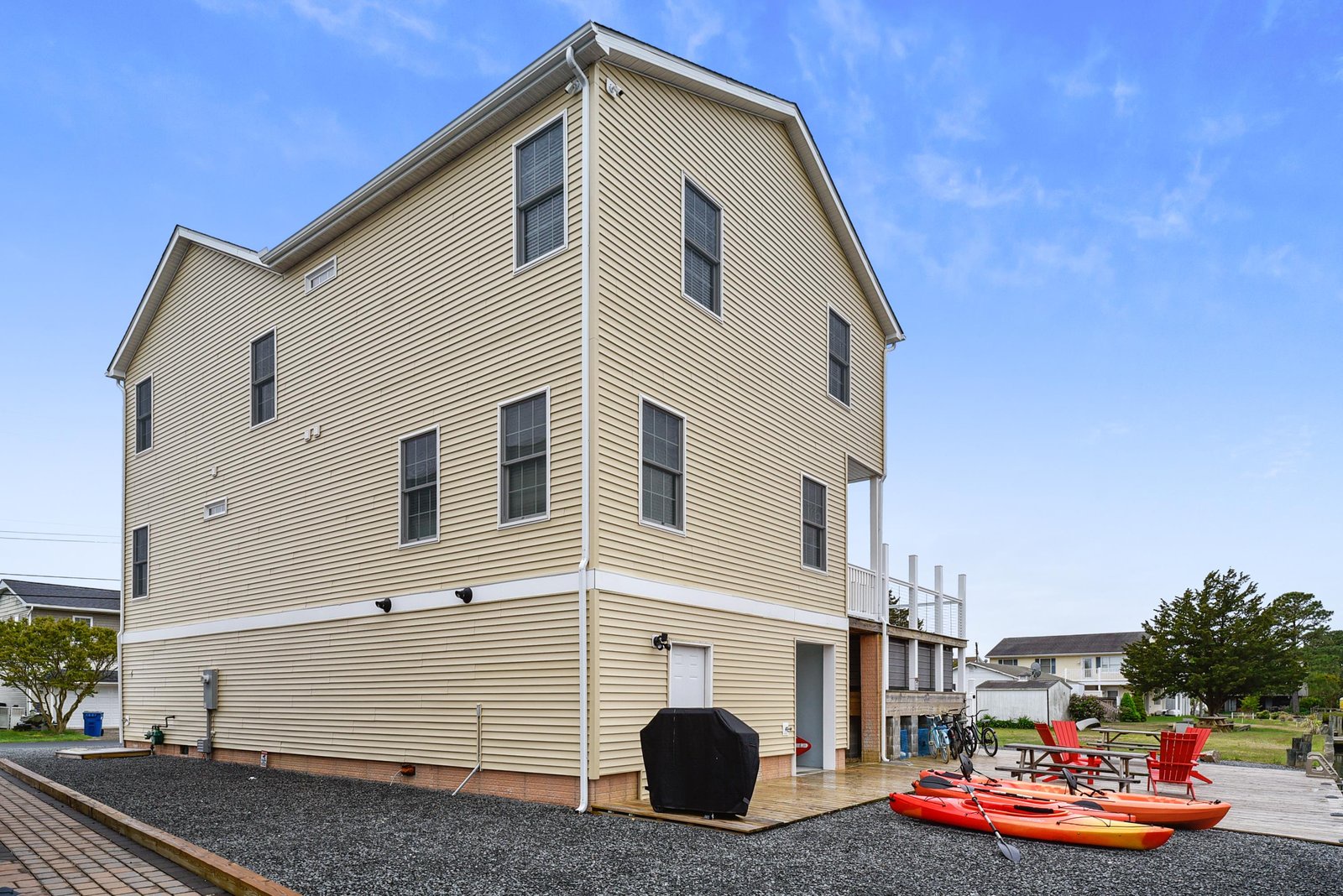 A spacious three-story coastal property with multiple levels of outdoor spaces, including a ground-level deck with colorful seating and kayaks ready for your waterfront adventures.