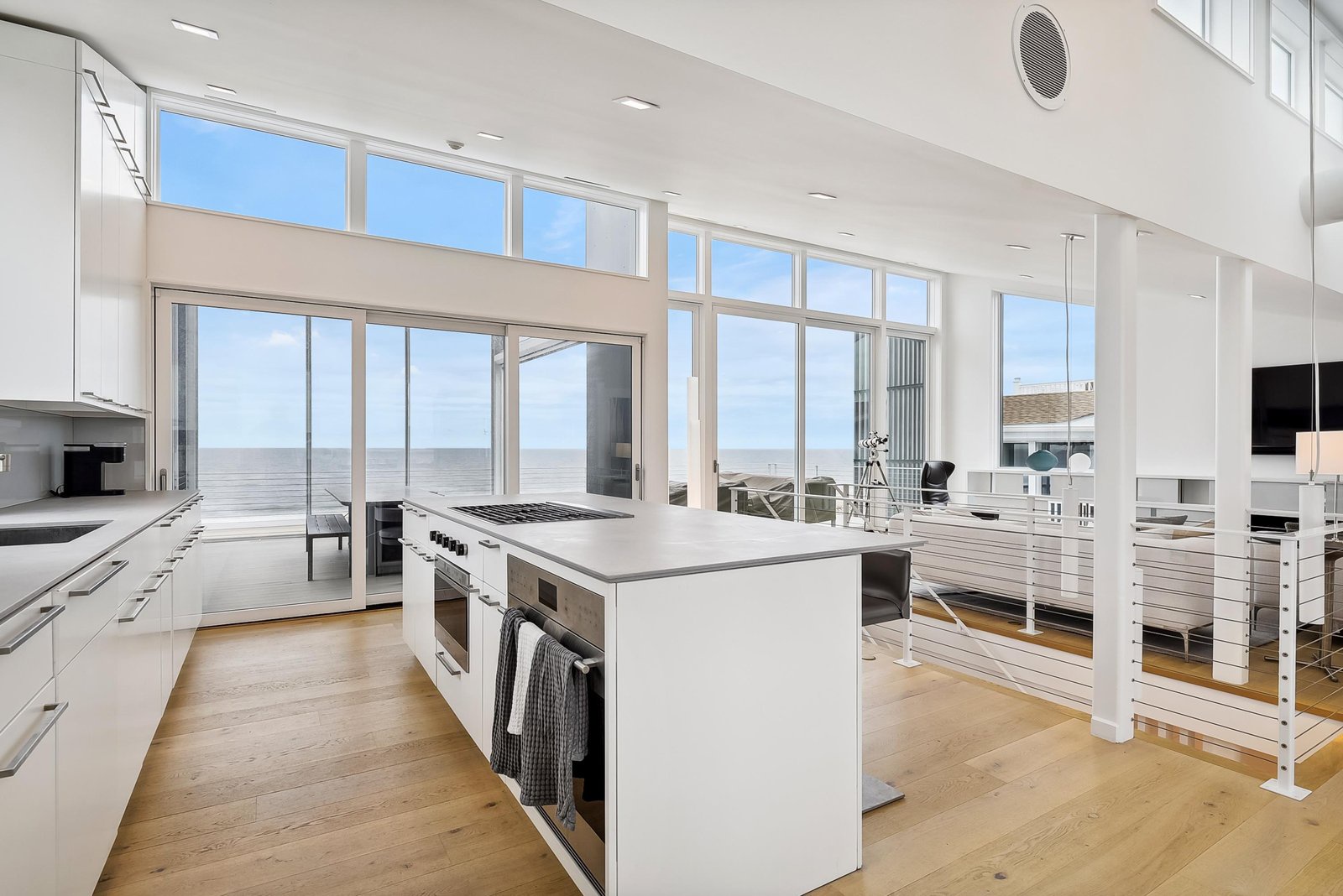 Step into your bright coastal kitchen where soaring ceilings, sleek white counters, and floor-to-ceiling windows frame stunning water views—your perfect backdrop for morning coffee or preparing a memorable meal.