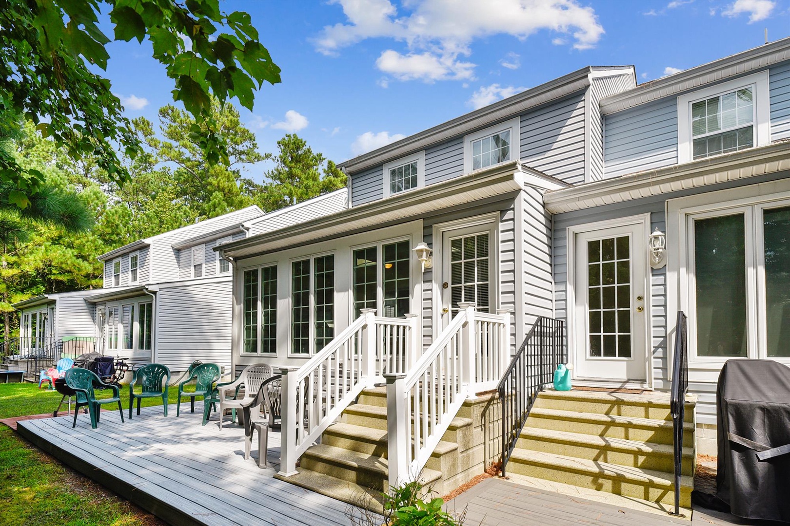 A charming property exterior featuring classic gray siding, white trim, and multiple entrances. The backyard deck offers outdoor seating under mature trees, ideal for relaxing and outdoor dining during warm, sunny days.