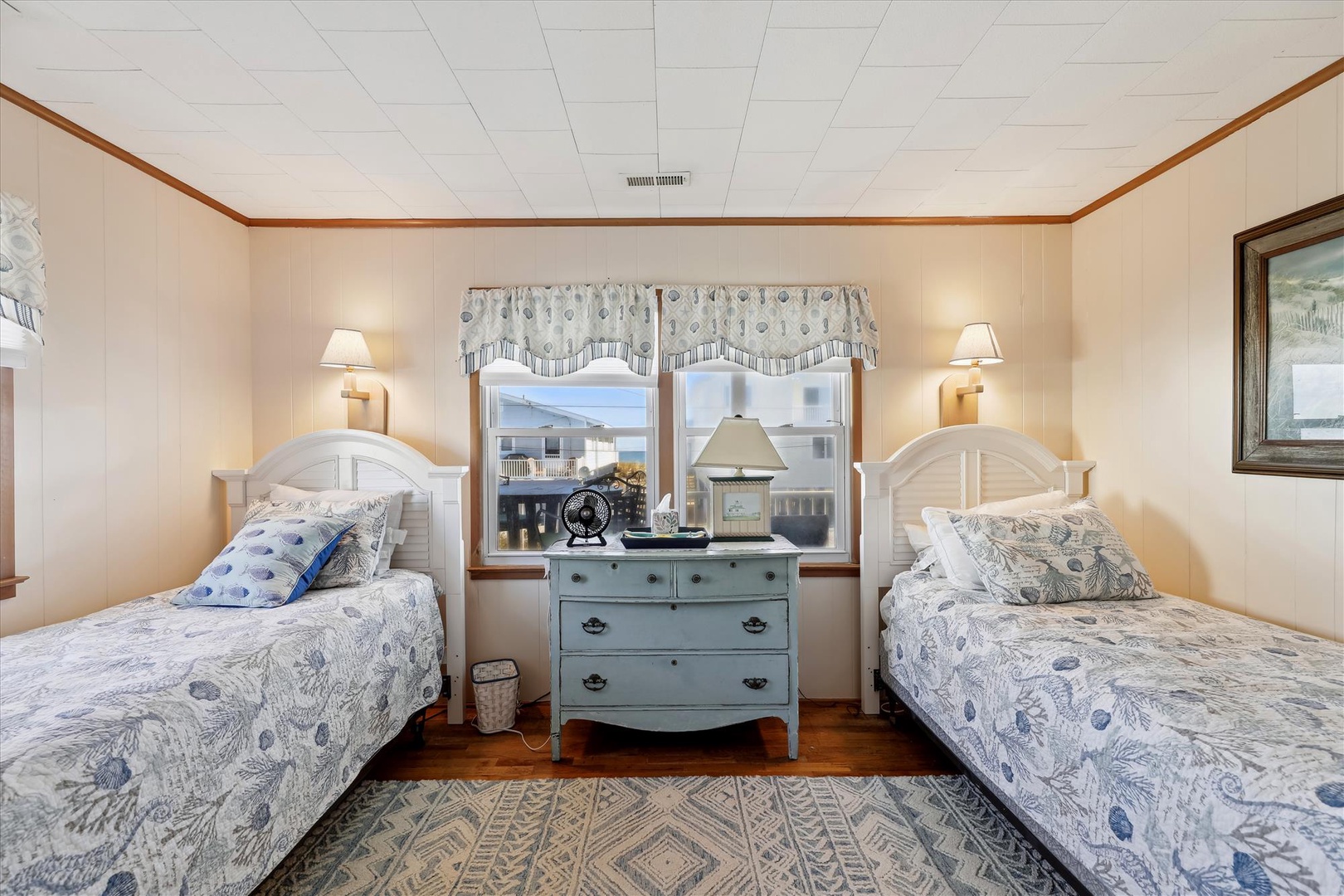 Two cozy twin beds with coastal-inspired linens frame a charming vintage dresser, perfect for your group getaway. Natural light pours in as you unwind in this welcoming retreat.