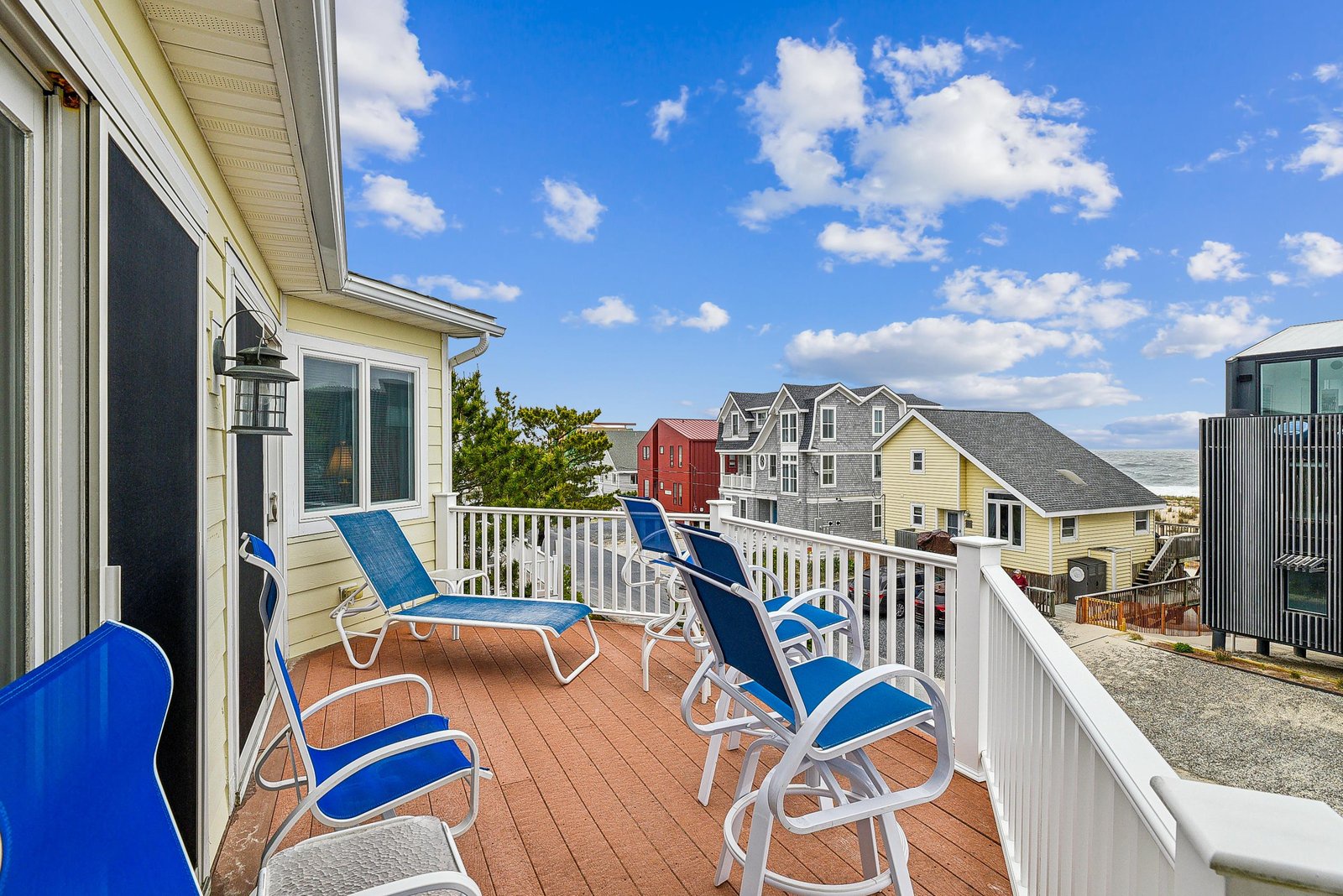 A spacious upper-level balcony with comfortable outdoor seating overlooks a quiet coastal neighborhood, offering a pleasant spot for morning coffee or evening relaxation under open skies.