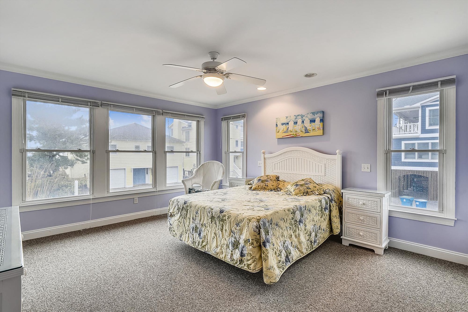 Unwind in this serene bedroom featuring a plush queen bed, calming lavender walls, and abundant natural light from multiple windows. The coastal artwork and white furnishings create your perfect retreat.