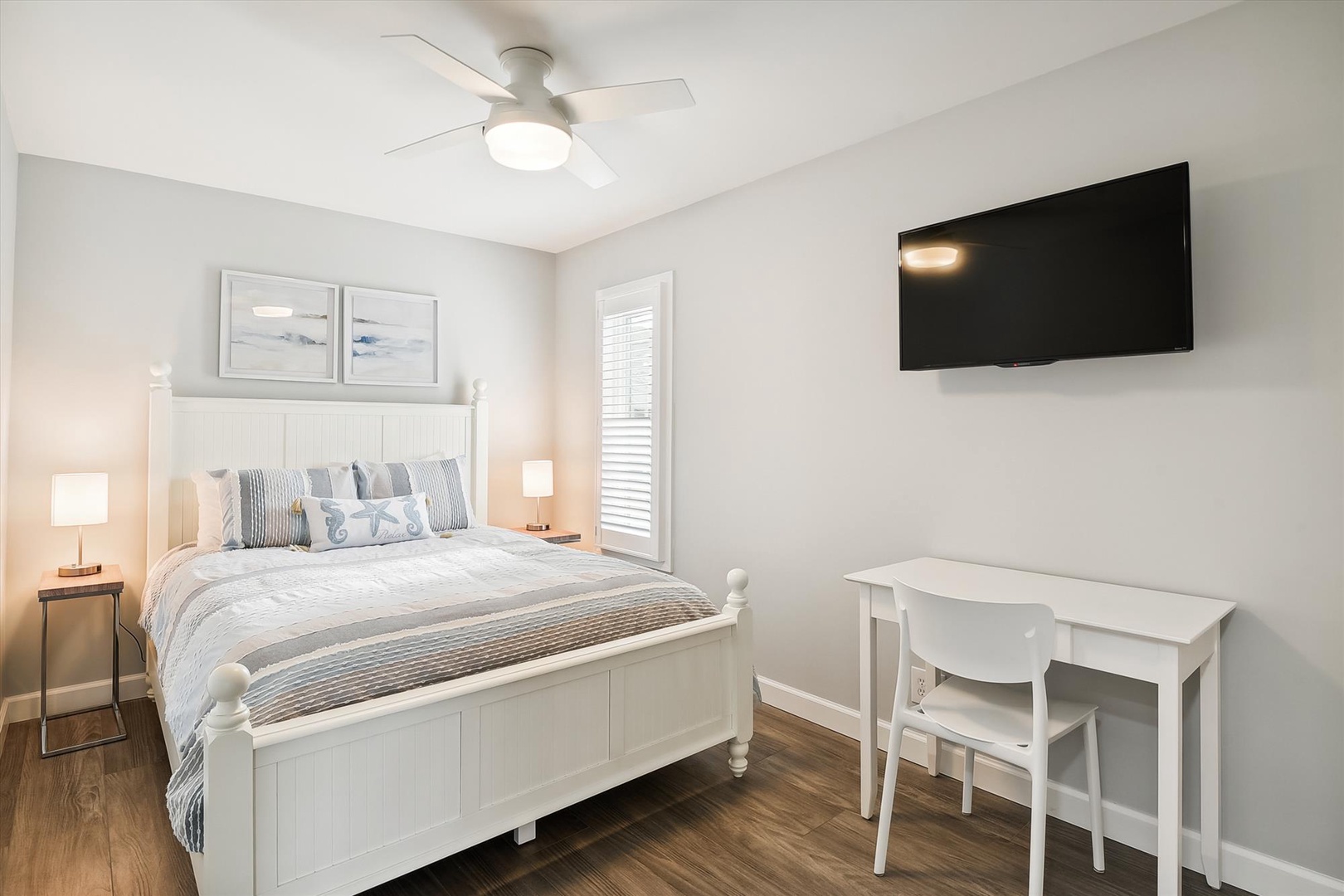 Your coastal-inspired bedroom offers a peaceful retreat with a plush bed dressed in soothing neutrals, a mounted TV for evening relaxation, and a convenient workspace by the window for morning coffee or planning your day.