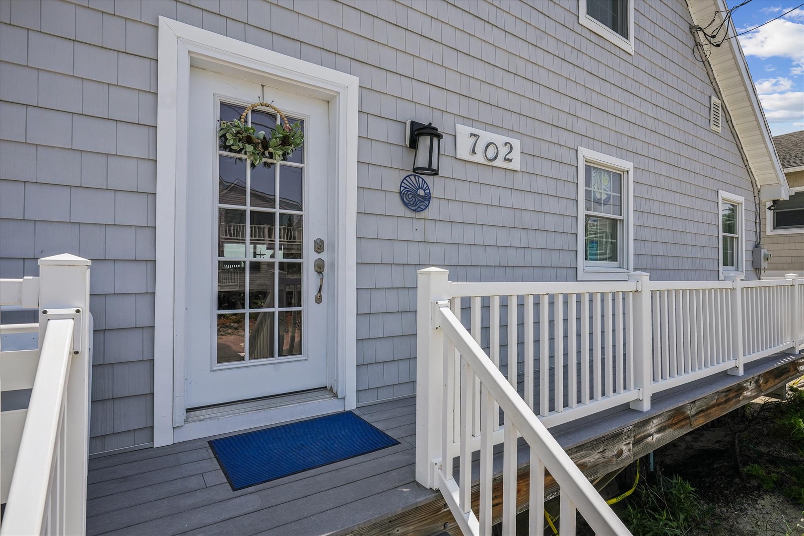 Property entrance featuring gray shingled exterior with white trim, decorative wreath, and address number 702. Small covered porch with white railing marks the arrival point to this coastal-style vacation home.