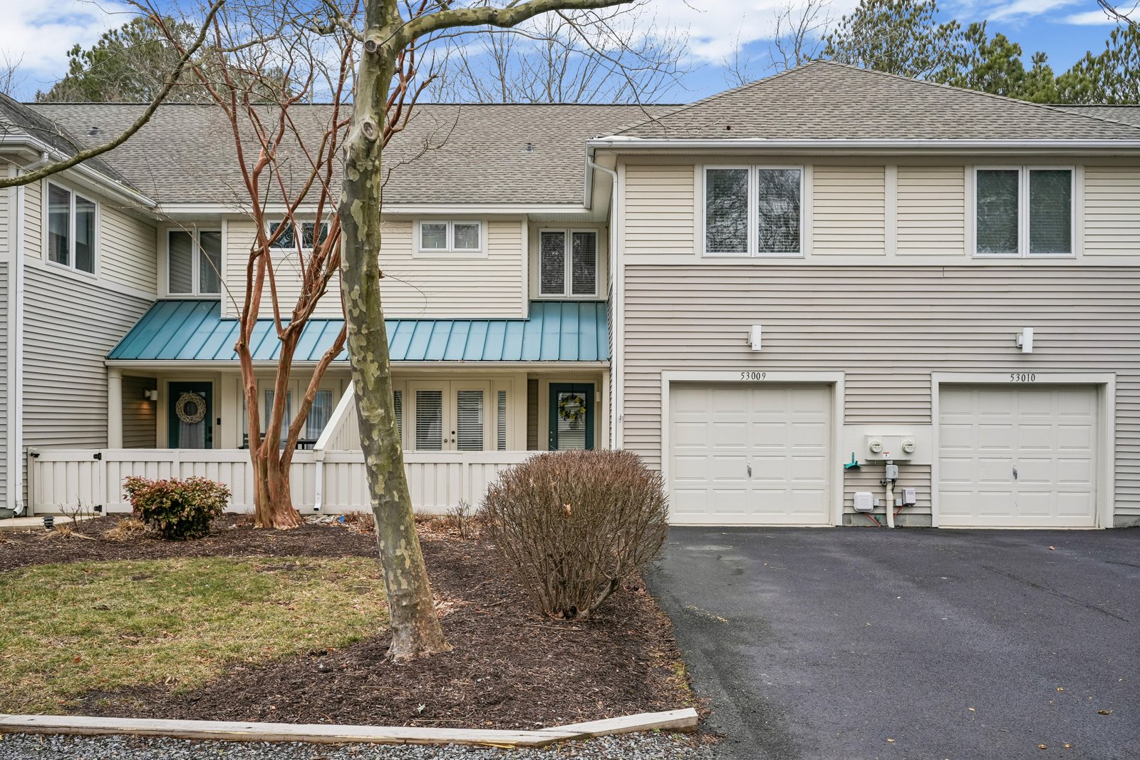 A modern two-story duplex with turquoise metal awnings, white garage doors, and address markers, situated in a wooded neighborhood with mature trees and convenient parking.