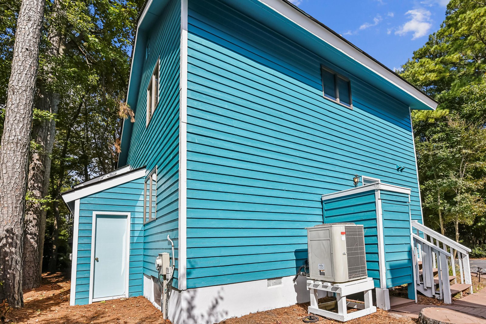 A bright turquoise two-story vacation home tucked among tall trees, offering a peaceful woodland retreat with modern comforts and private outdoor space.