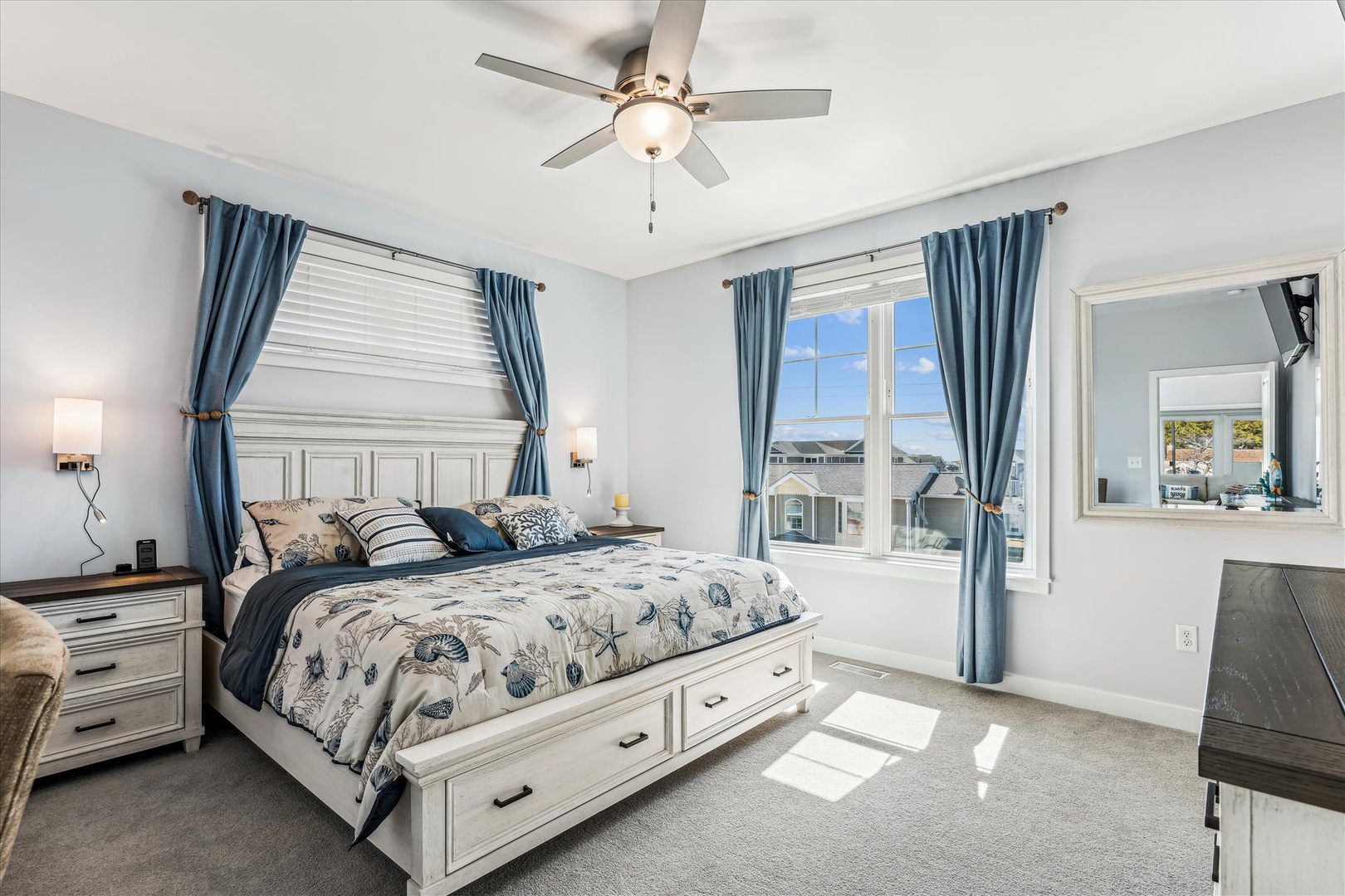 Relax in your bright, coastal-styled bedroom with ocean-themed bedding and gentle breezes from the ceiling fan. Blue curtains frame windows that fill the space with natural light.
