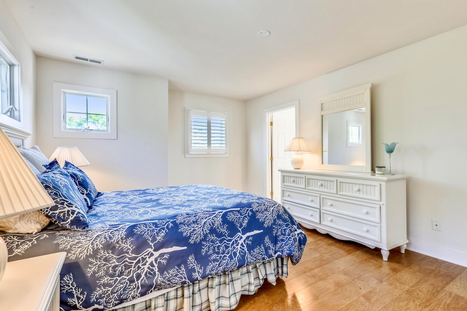 Settle into this cozy bedroom with crisp white furnishings, warm wood floors, and soft coastal-blue linens. Natural light from the windows creates a serene retreat for restful nights.
