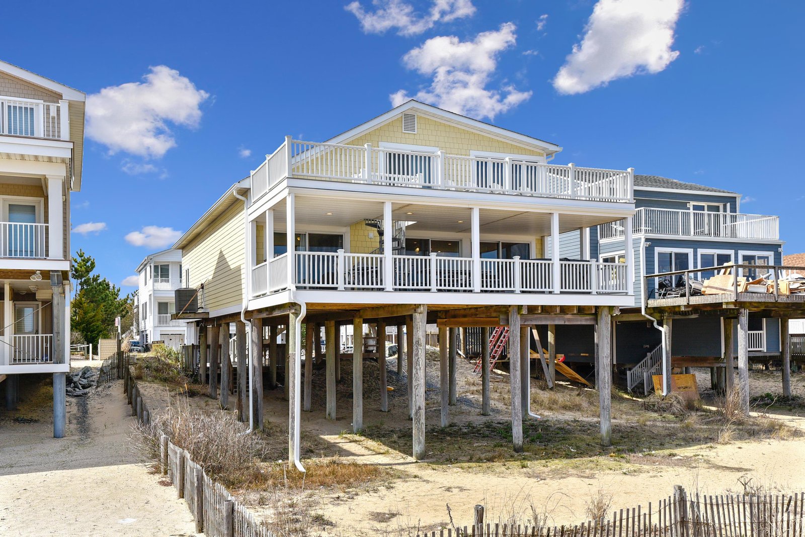 A coastal beach house elevated on stilts with spacious decks and balconies, situated in a residential beachfront neighborhood under a bright blue sky.