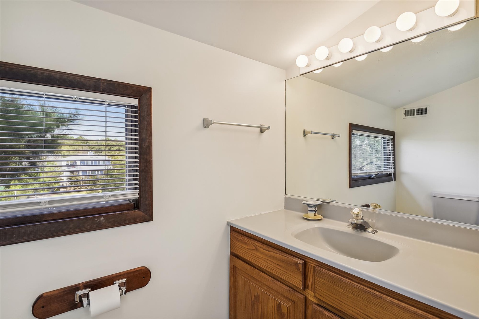 Your private bathroom awaits with warm wood cabinetry, bright vanity lighting, and a sunny window that fills the space with natural light.
