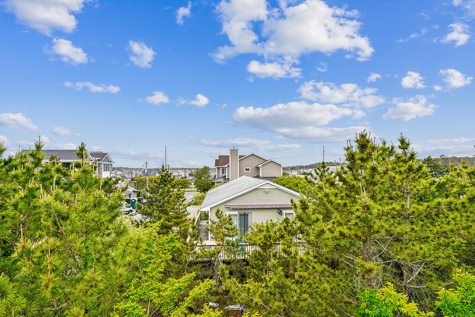 The property sits nestled in a coastal neighborhood, surrounded by lush pine trees and under a brilliant blue summer sky, offering a peaceful and scenic backdrop for your stay.