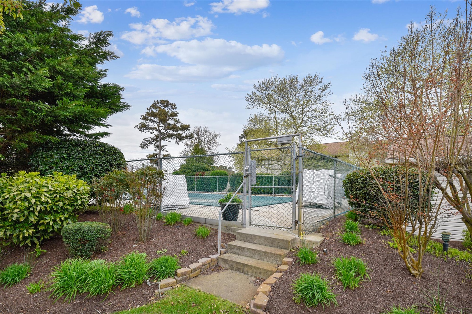The property features a tennis court surrounded by mature trees and landscaped gardens, accessible via gated entry and steps through the grounds.