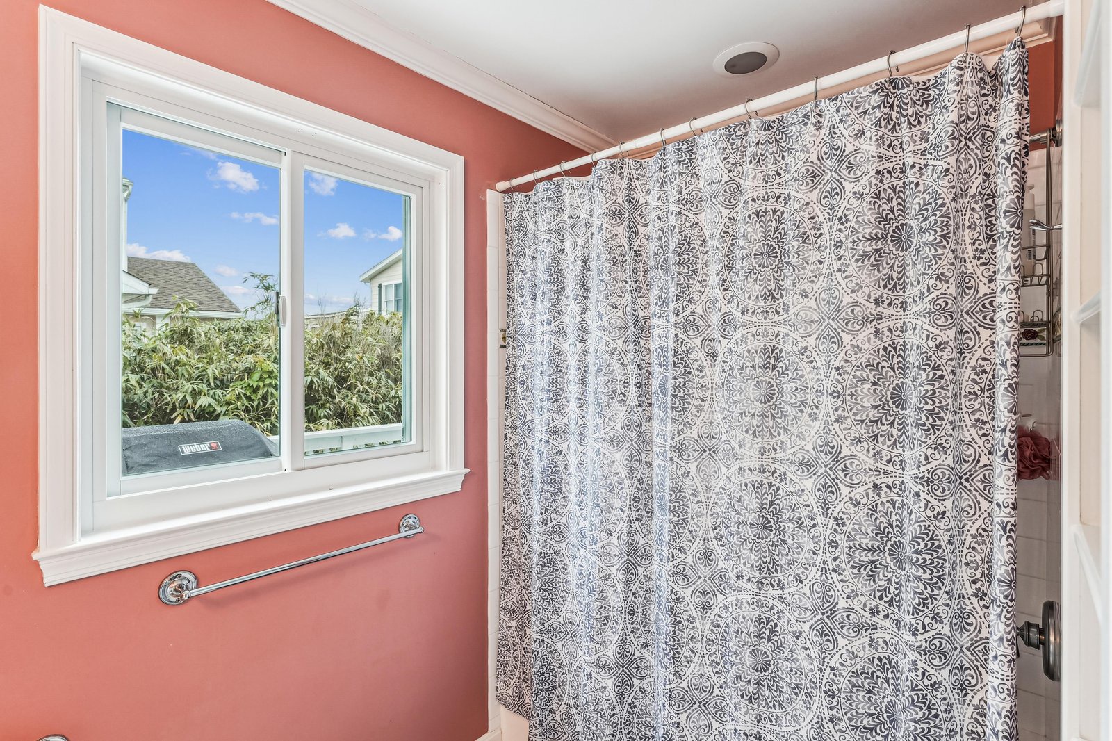 Step into your charming coral-toned bathroom with a patterned shower curtain and natural light streaming through the window overlooking the garden. It's the perfect spot to freshen up and start your day.